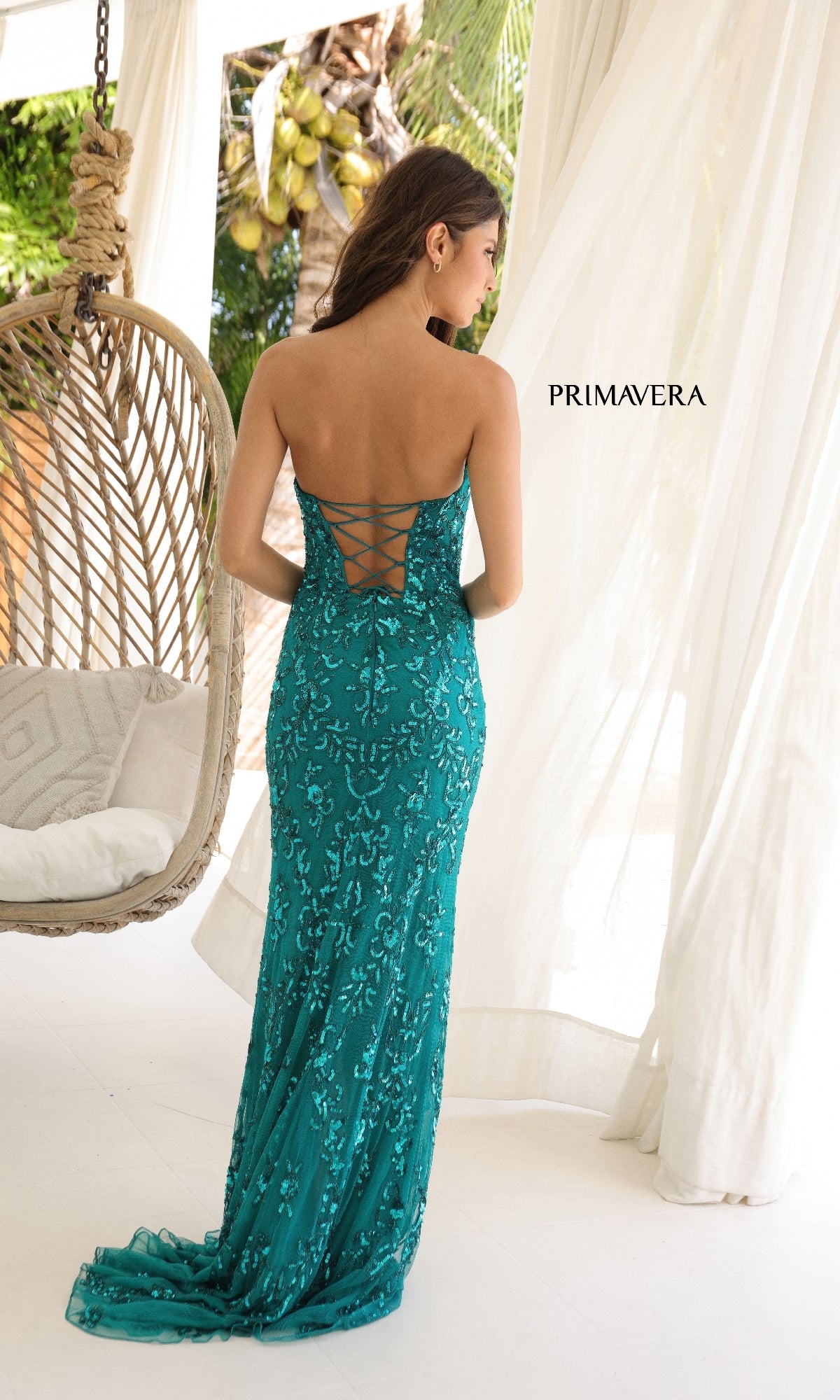A woman in a teal Primavera 4347 strapless long sequin prom dress with lace-up back stands indoors near a hanging chair and sheer white curtains.
